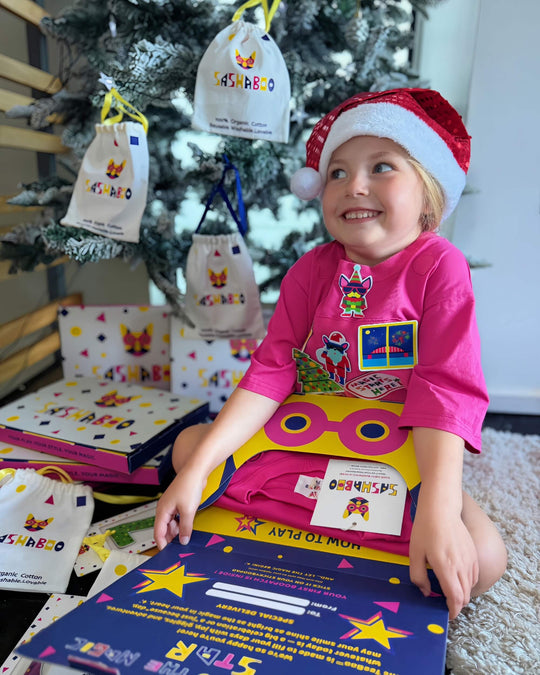 Child wearing a pink TeeBoo™ interactive T-shirt with Christmas BooPatch™ designs beside SashaBoo gift packaging and play activities