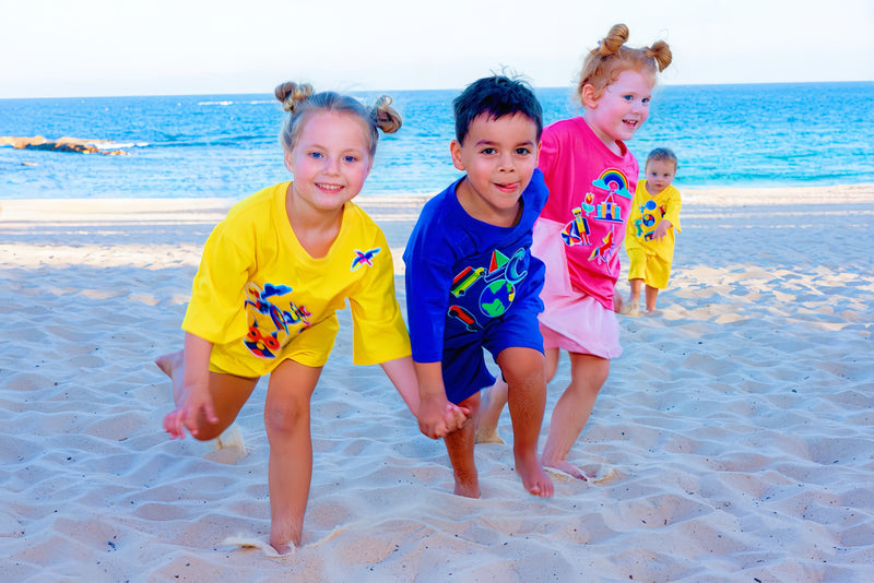Three children playing on a sandy beach wearing colourful SASHABOO interactive TeeBoo T-shirts with interchangeable BooPatch designs, with the ocean in the background.