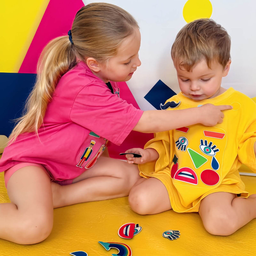 Two children playing together with SASHABOO Moody Friends interactive T-shirts. A girl points to colourful interchangeable emotion patches on a boy's yellow TeeBoo T-shirt.